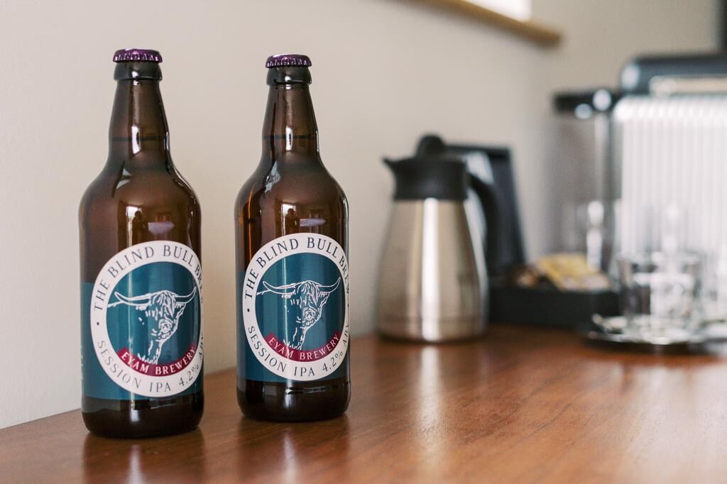 Duas garrafas de The Blind Bull Session IPA 4.2% sobre uma mesa de madeira, com uma cafeteira e copos ao fundo.