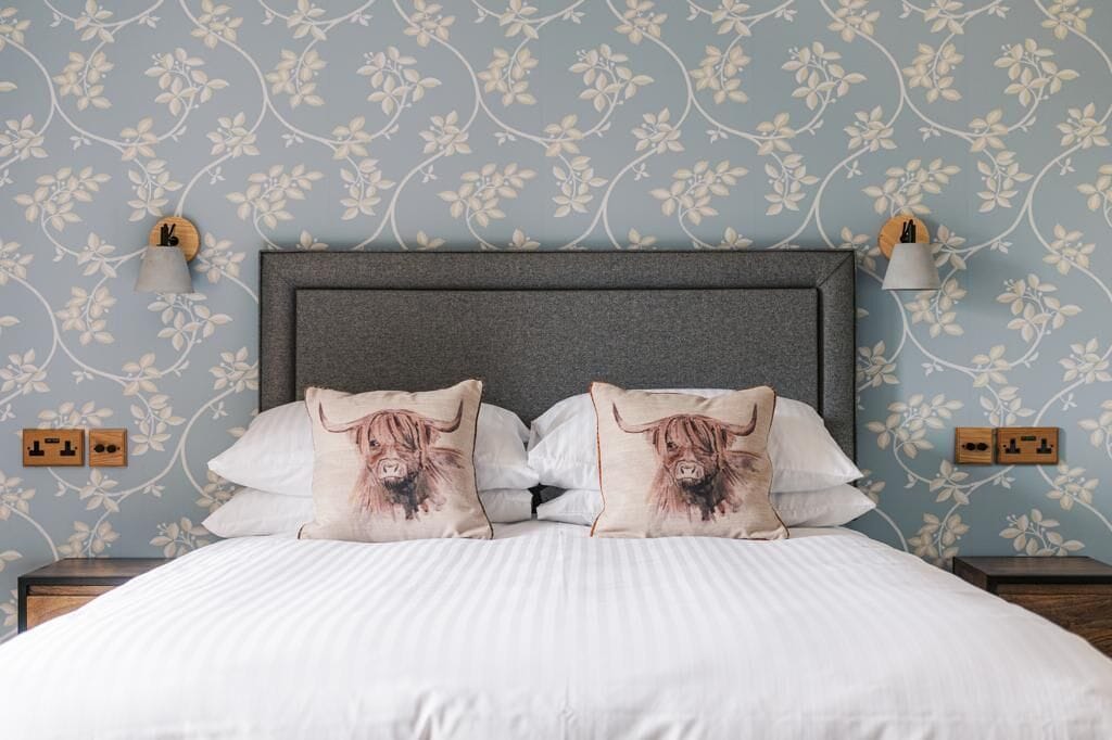 Una cama bien tendida con ropa blanca y dos almohadas con un diseño de vaca de las Tierras Altas, sobre un fondo de papel tapiz floral.