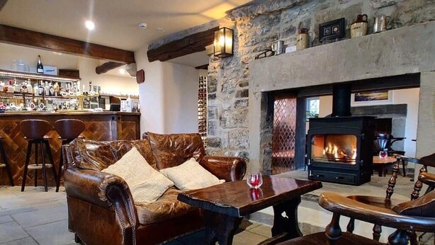 A cozy living room with a stone fireplace, leather sofa, wooden table, and bar area with stools. - The Blind Bull (Buxton)