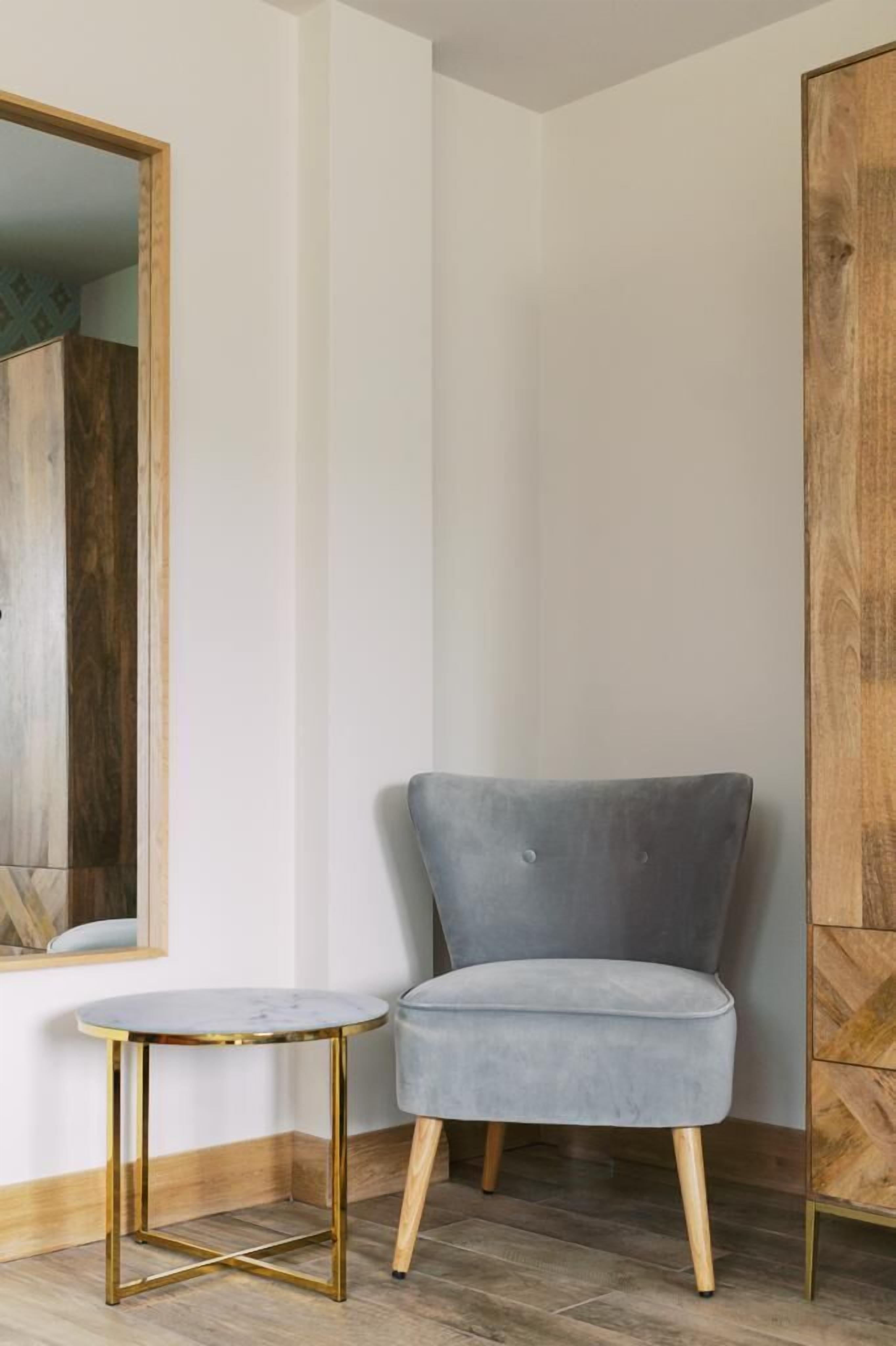 Una habitación de esquina con un sillón tapizado gris, una mesita redonda con marco dorado y un aparador de madera.