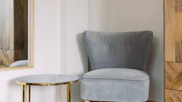 Una habitación de esquina con un sillón tapizado gris, una mesita redonda con marco dorado y un aparador de madera.