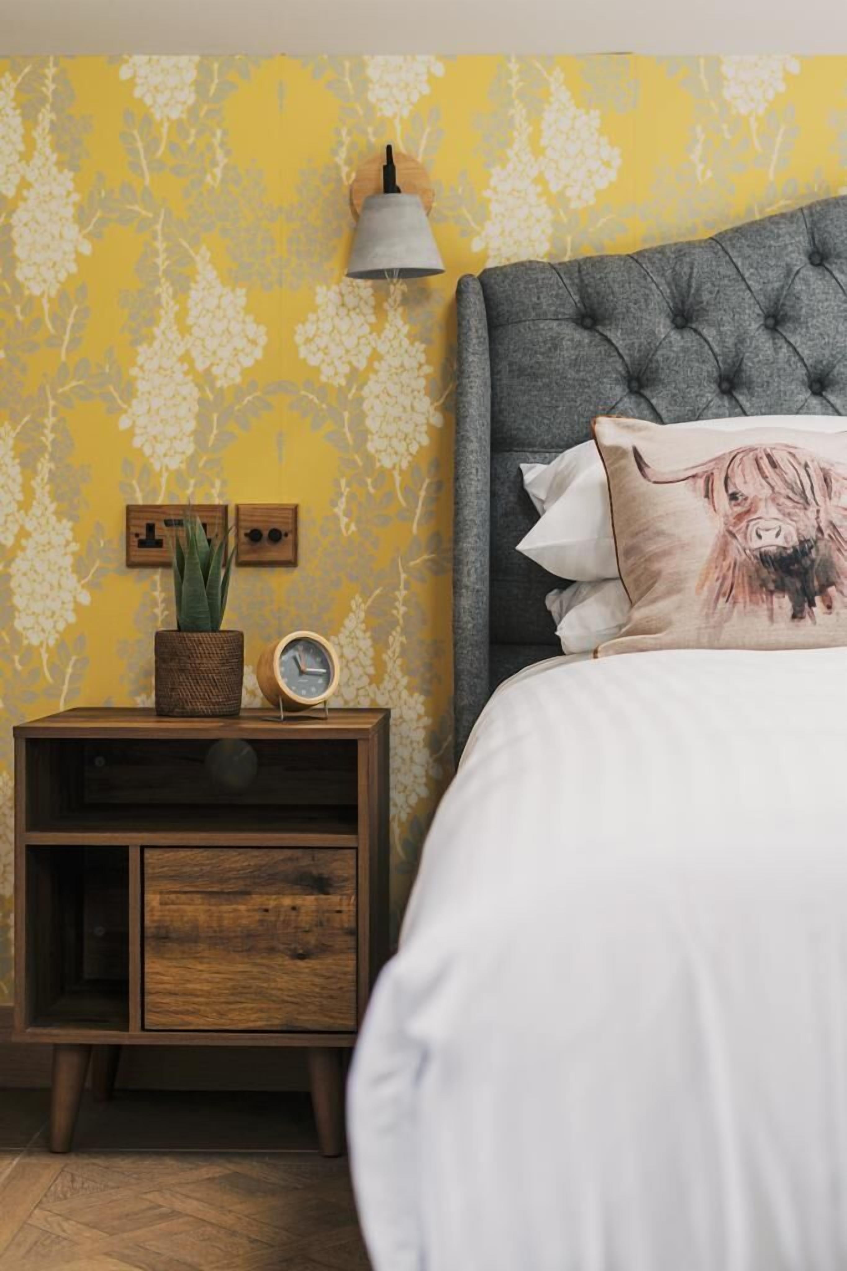 A bedroom with a tufted headboard, a bedside table with a clock and a plant, and a wall with floral wallpaper.