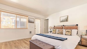 Un dormitorio con una cama grande, piso de madera, un banco y una ventana con persianas.
