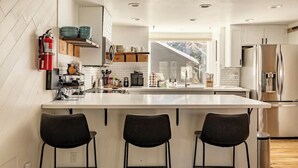 Una cocina moderna con una isla grande, muebles blancos y electrodomésticos de acero inoxidable. Hay tres taburetes negros y un extintor de incendios montado en la pared.