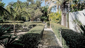 Un chemin bordé de haies et de palmiers soigneusement taillés, qui mène à un bâtiment blanc.