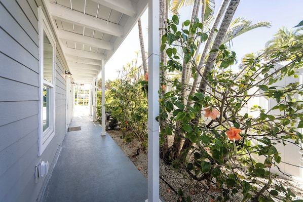 Eine überdachte Veranda mit Betonboden, weißer Außenverkleidung und Blick auf einen Garten mit verschiedenen Pflanzen und einer Hibiskusblüte.