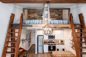 Et kompakt køkken med en central ø, et køleskab og en mikrobølgeovn. Der er træstiger på hver side af køkkenet, og et loftsrum med soveplads og en hængende lampe over sengen.