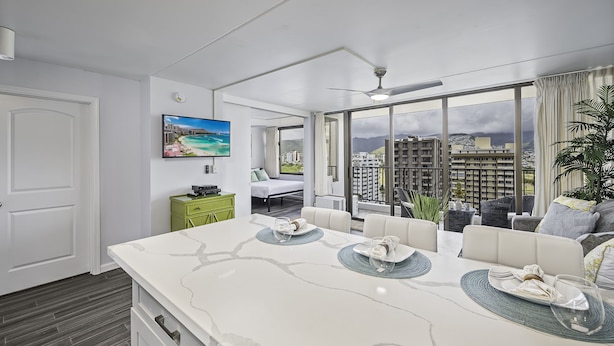 A modern hotel room with a large dining table, a bed, and a balcony with a city view. - City-Golf Course View Condo at the Waikiki Banyan w/Free Parking & WiFi (Honolulu)