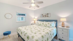 A bedroom with a bed, a ceiling fan, a nightstand with a lamp, a small stool, and a window with a view of a building. - Mid-Island, completely renovated duplex, 1bedroom/1 bath- CLOSE TO THE BEACH! (Fort Myers Beach)
