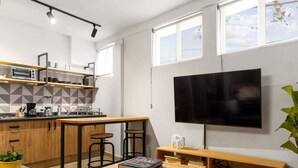 A modern kitchen with a central island, a dining area with a wooden table and chairs, a flat-screen TV mounted on the wall, and a large window providing natural light. - MS Moderna Suite Arcos Hercules (Santiago de Querétaro)