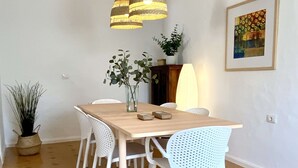 A dining area with a wooden table, white chairs, hanging pendant lights, a potted plant, and a framed picture on the wall.