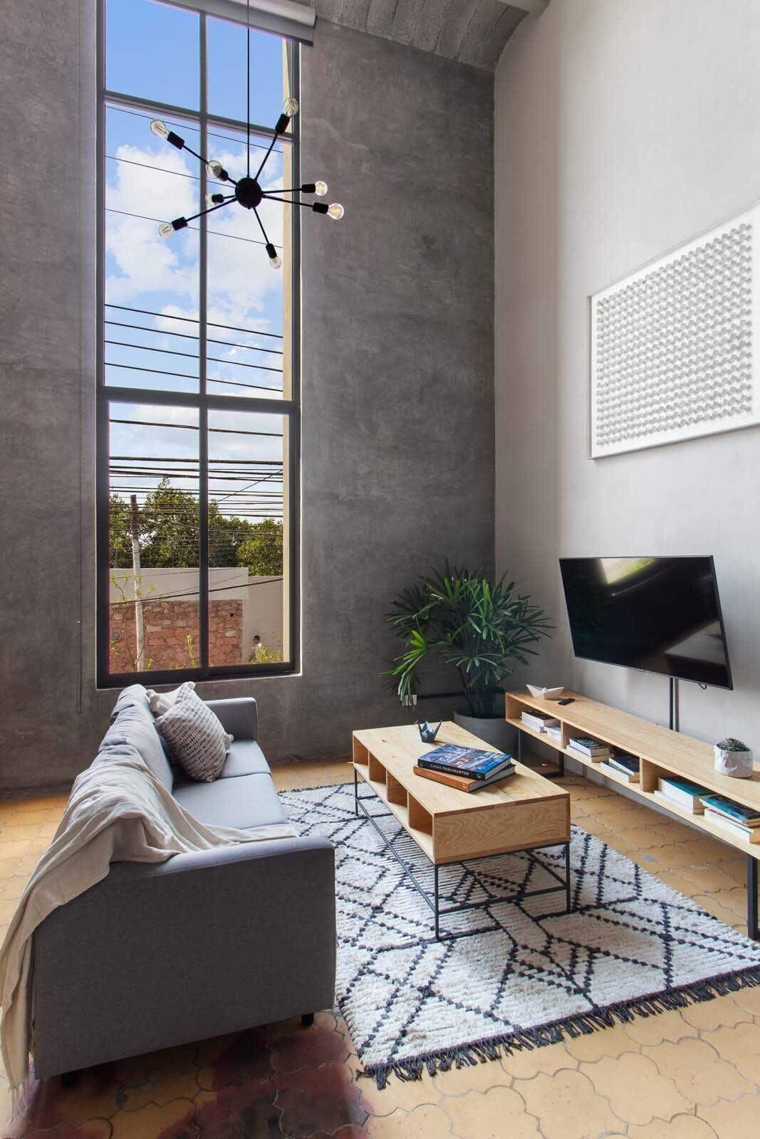 Un salon moderne avec une grande fenêtre, un canapé gris, une table basse en bois, un téléviseur et un tapis à motifs.