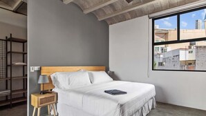 A bedroom with a bed, a bedside table with a lamp, a window with a view of buildings, and a concrete floor. - +MS +Contemporáneo loft +Ubicadísimo +Arcos (Santiago de Querétaro)