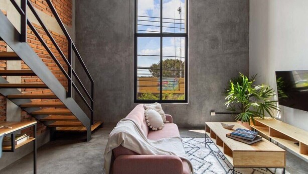 A modern living room with a brick wall, a large window, a staircase, a sofa, a TV, and a plant. - +MS +Contemporáneo loft +Ubicadísimo +Arcos (Santiago de Querétaro)