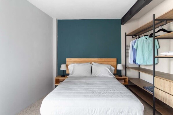 Une chambre à coucher comprenant un lit, deux tables de chevet, une étagère murale avec des vêtements et un mur d’accent de couleur turquoise.