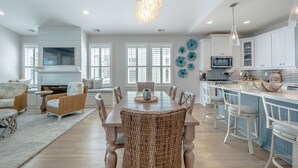 A spacious living room with a dining area, kitchen, and a large window with shutters. - Serenity by the Sea | Community Pool + Near Beach! (Kure Beach)