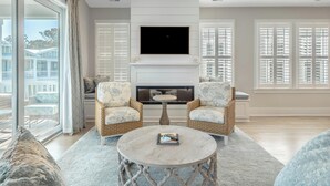 A spacious living room with a fireplace, two wicker armchairs, a round coffee table, and large windows offering a view of a residential area. - Serenity by the Sea | Community Pool + Near Beach! (Kure Beach)