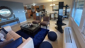 Una sala moderna con un sofá, sillones, una mesa de centro de vidrio y un televisor. Se observa una cocina al fondo, con una zona de comedor.