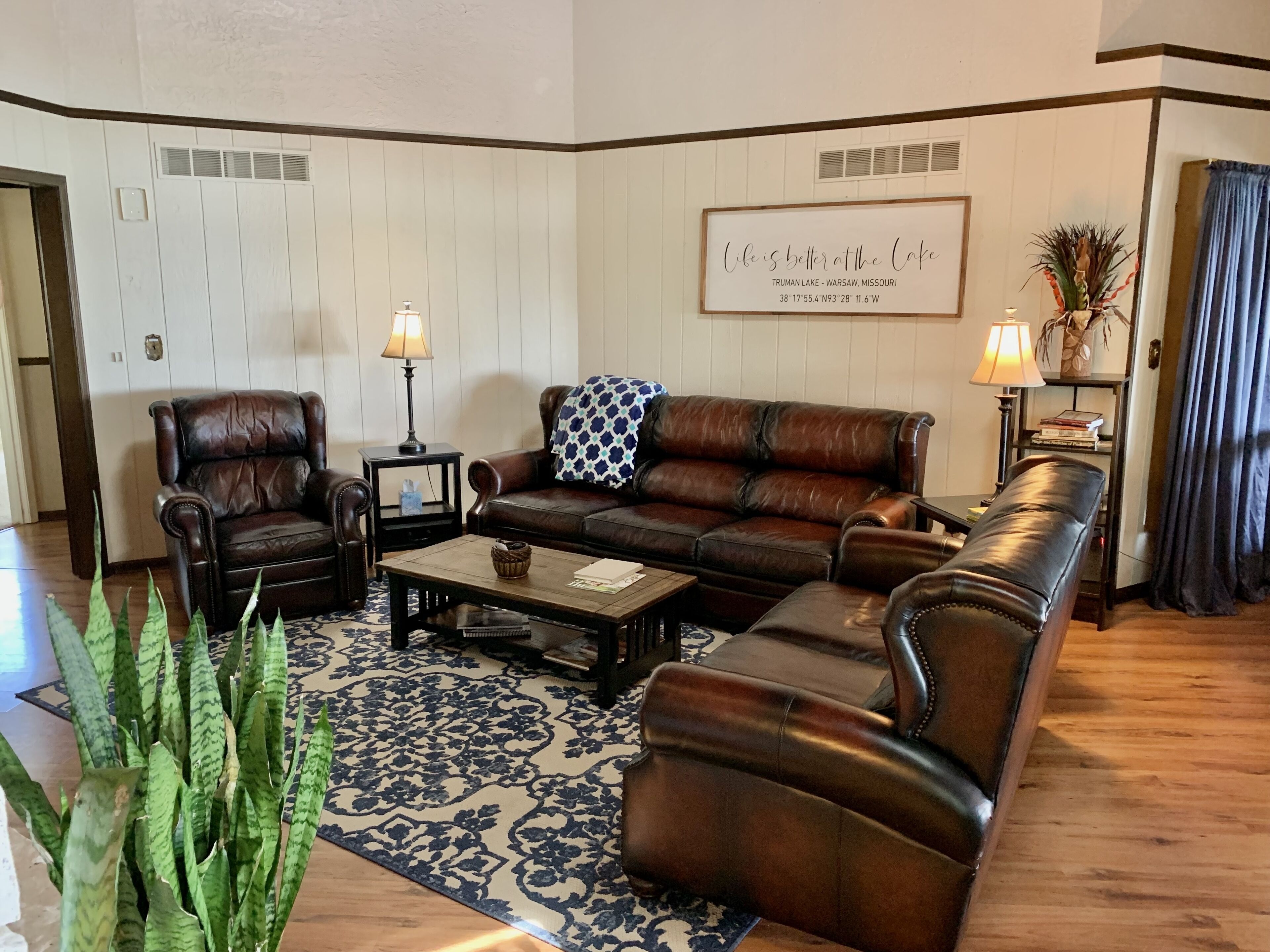 A living room with a brown leather sofa, a matching armchair, a coffee table, and a patterned rug. There is a framed sign on the wall and a potted plant in the corner.
