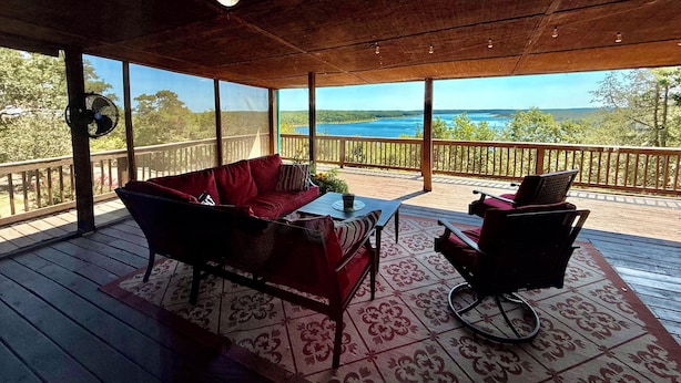 Un espacio de estar al aire libre cubierto con un sofá rojo y una mesa, con vista a un paisaje pintoresco de un lago y un bosque.