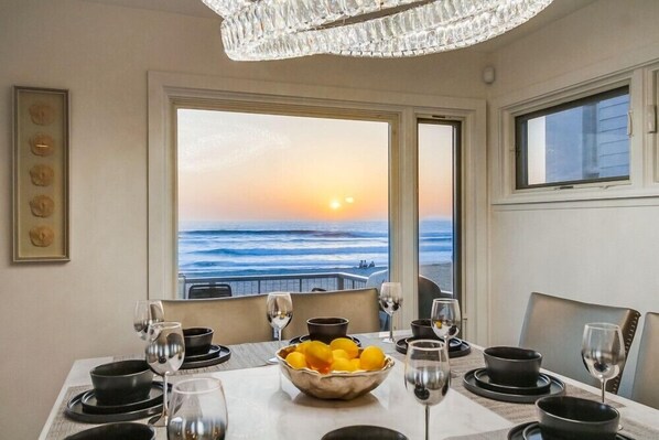 Un comedor con una mesa dispuesta para cuatro, un frutero y vistas al océano al atardecer.