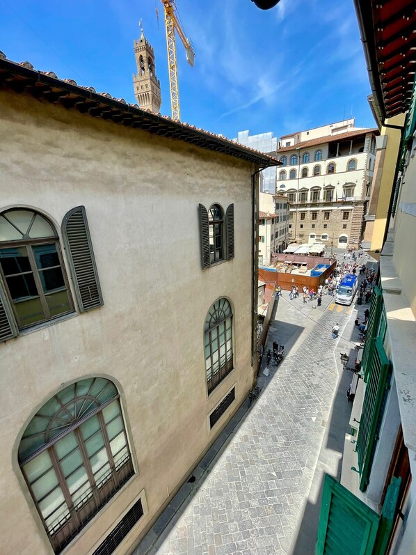 A narrow street with buildings on either side, a market area with people and stalls, and a tower in the background.