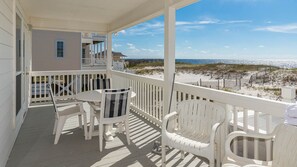 Un espace extérieur abrité, doté de mobilier blanc, comprenant des chaises et une table, et offrant une vue sur une plage avec des dunes de sable et une clôture en bois.