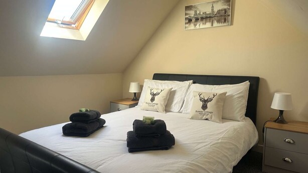 A neatly made bed with white linens and two pillows featuring a deer logo, a bedside table with a lamp, and a framed cityscape picture on the wall. - Cottage with Log Burner - Wanlockhead D & G (Wanlockhead)