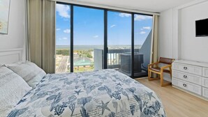 Une chambre à coucher avec un grand lit, une chaise, une commode et une vue sur le paysage urbain, visible à travers de grandes fenêtres.