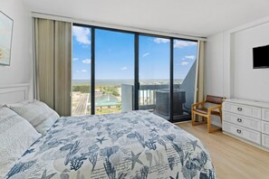 Une chambre à coucher avec un grand lit, une chaise, une commode et une vue sur le paysage urbain, visible à travers de grandes fenêtres.
