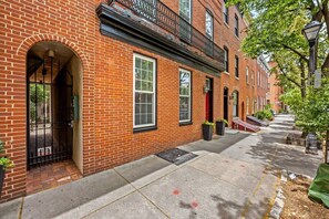 A brick building with a red door, a black metal gate, and a sidewalk with a green planter.