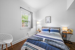 A bedroom with a bed, a chair, a bedside table, a lamp, and a window with curtains.