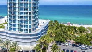 Un immeuble de grande hauteur à la façade incurvée, un parking rempli de voitures et, au premier plan, une plage bordée de palmiers.