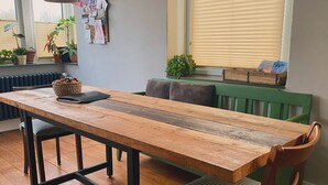 A dining area with a wooden table, black metal frame, and two chairs. There are hanging lights, a radiator, and potted plants on the windowsill.
