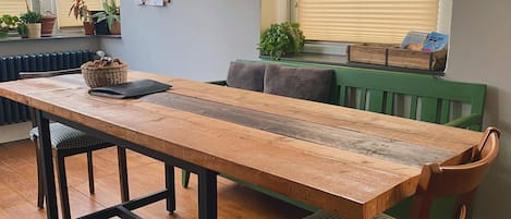 Ein Essbereich mit Holztisch, schwarzem Metallgestell und zwei Stühlen. Es gibt Pendelleuchten, eine Heizung und Blumentöpfe auf der Fensterbank.