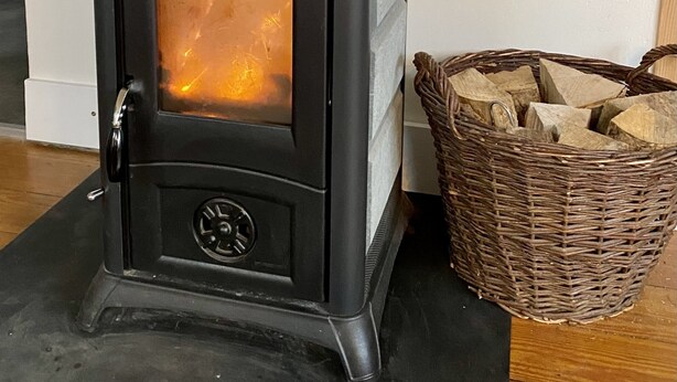 A wood-burning stove with a glass door, a black chimney, and a wicker basket of firewood beside it.