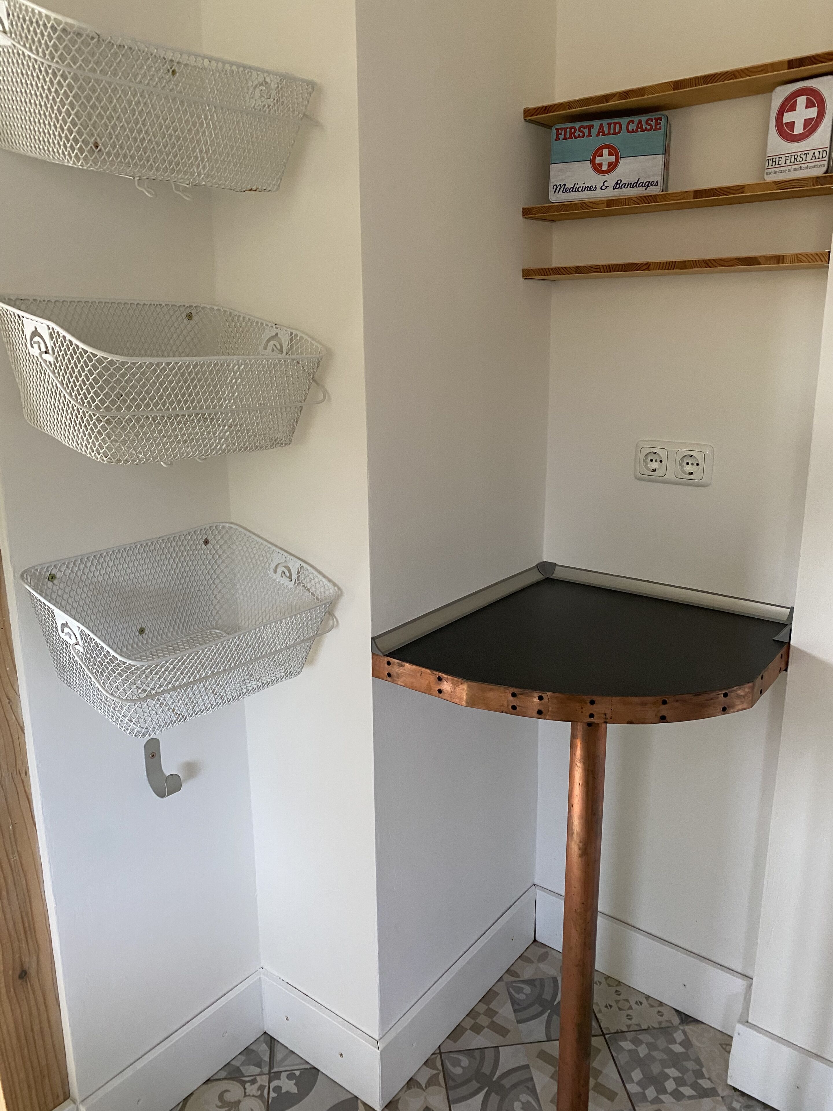 A corner with a metal basket shelf, a small table with a black top, and a shelf with a first aid kit and a box labeled "Medicines & Bandages".