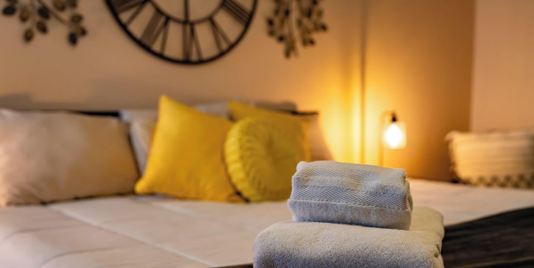A bed with white bedding and two rolled towels, a clock on the wall, and a bedside lamp.