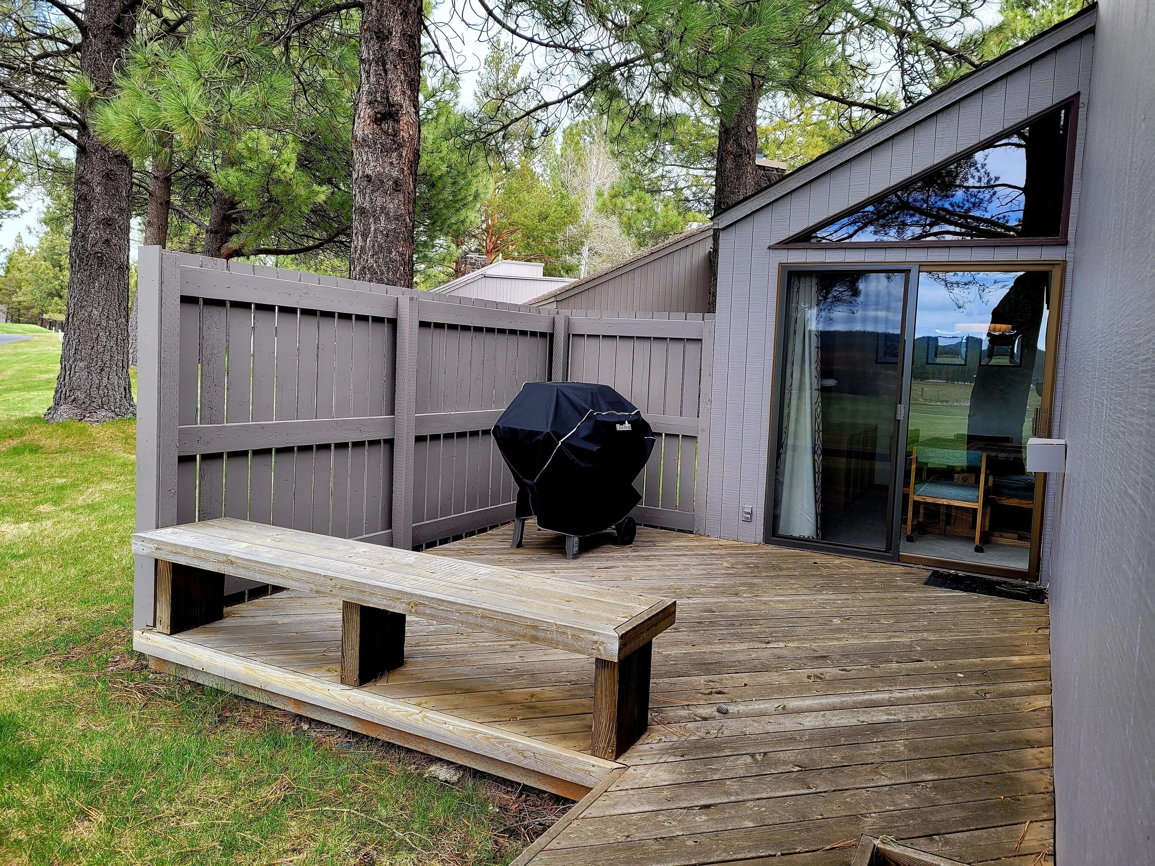 Een houten terras met een barbecue en een bankje, een glazen deur die leidt naar een overdekt gedeelte en een houten omheining.