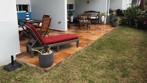 A covered patio with a red lounge chair, a wooden table, and chairs. There are potted plants and a small garden area. - 3 Bedrooms Apt. close to main beaches/ attractions (Río Grande)
