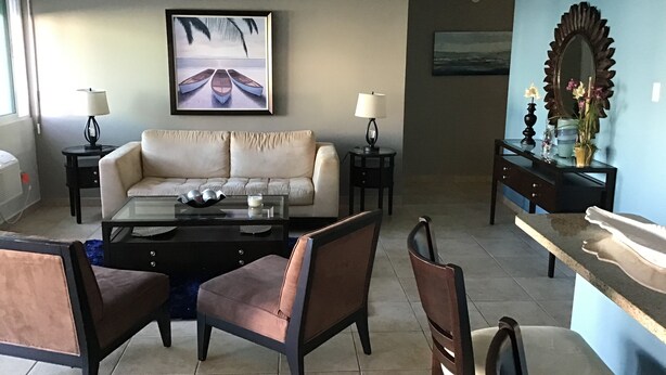 A living room with a sofa, two armchairs, a coffee table, and a dining area with a table and chairs. There are two lamps on side tables, a large painting above the sofa, and a mirror on the wall. - 3 Bedrooms Apt. close to main beaches/ attractions (Río Grande)