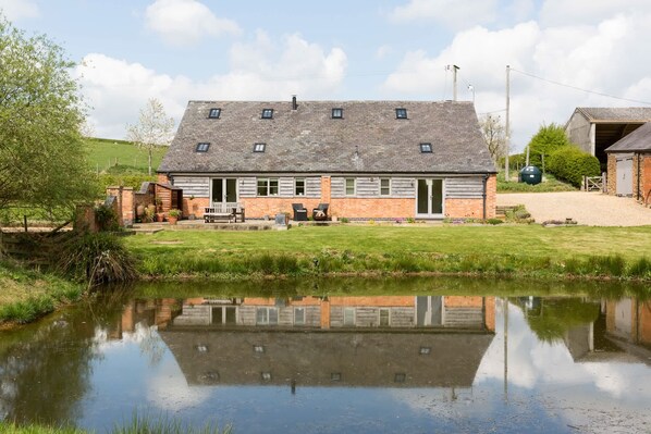 A two-story house with a sloped roof, a front porch with seating, and a small pond in the foreground. - Chater Barn (Leicester)