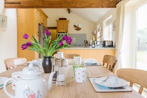 A dining table set with a vase of flowers, teapot, cups, and plates in a room with wooden furniture and a view of the kitchen. - Chater Barn (Leicester)