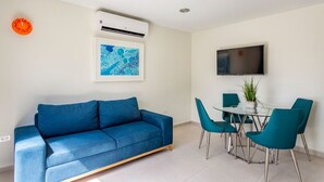 A modern living room with a blue sofa, a glass dining table with teal chairs, a mounted TV, and an air conditioning unit. - Keito 2BR APT #K3-3 min to Palm Beach-Sleeps 4 (Noord)