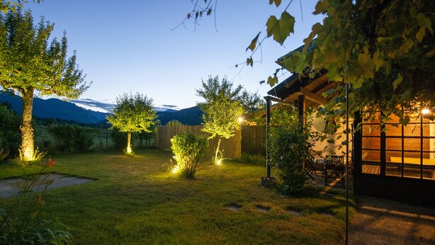 A well-lit backyard with a wooden pergola, a small table, and chairs under the shade of trees. - Zotlar House (Bohinj)
