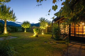 A well-lit backyard with a wooden pergola, a small table, and chairs under the shade of trees. - Zotlar House (Bohinj)