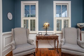 Una habitación con dos sillones, un escritorio de madera y dos ventanas con contraventanas.
