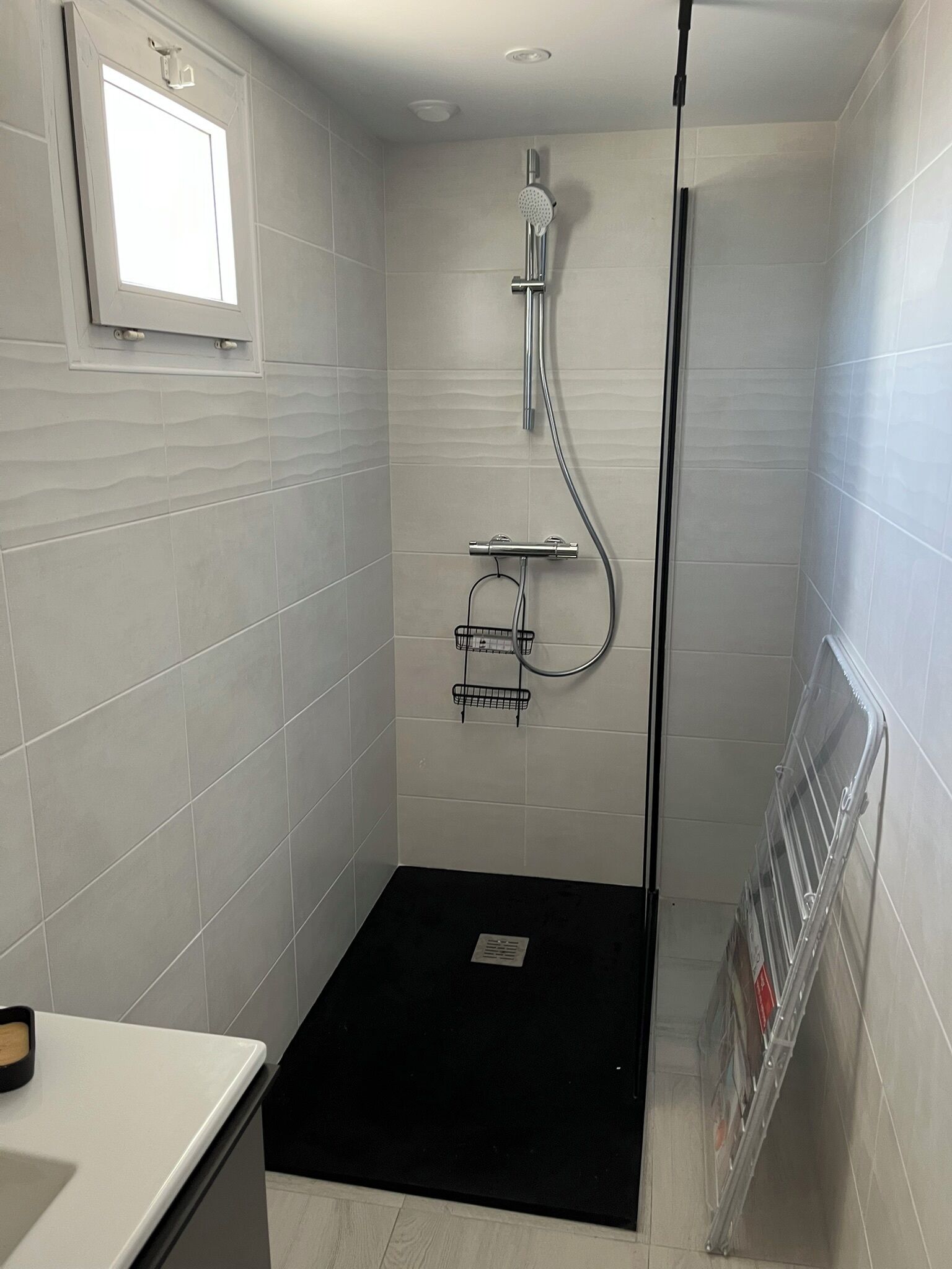 A modern bathroom with white tiles, a shower with a handheld showerhead, a black bath mat, and a small window.