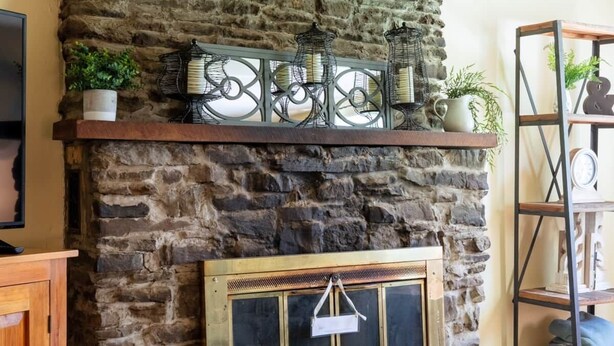 A stone fireplace with a metal screen door, a wooden mantel shelf with decorative items, and a wooden floor. - Dragonfly Cottage-Pet-friendly, relaxing Wellsboro retreat (Charleston Township)