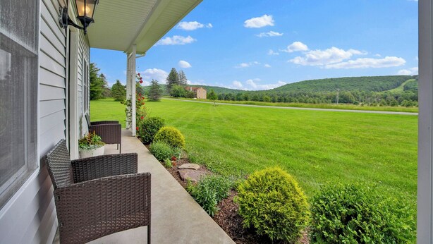 A porch with wicker chairs and a lamp, overlooking a green field and hills.